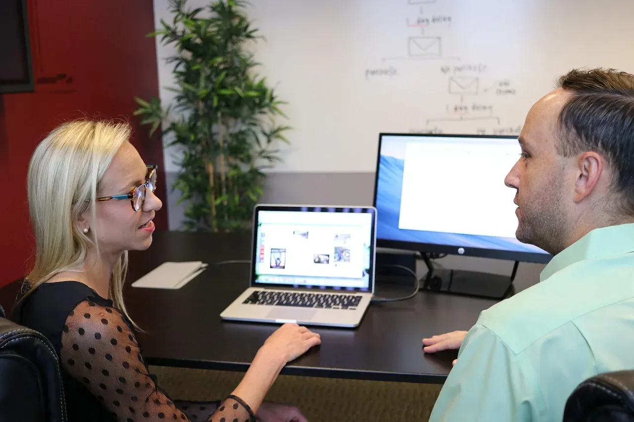 Two colleagues discussing work at a laptop and a desktop monitor