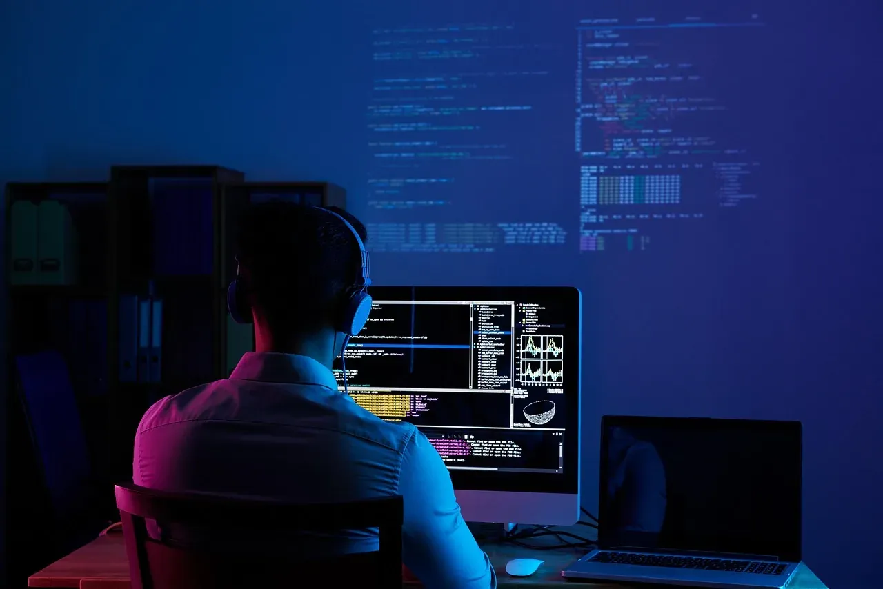 A developer in headphones working at a computer monitor, with code projected on the wall behind