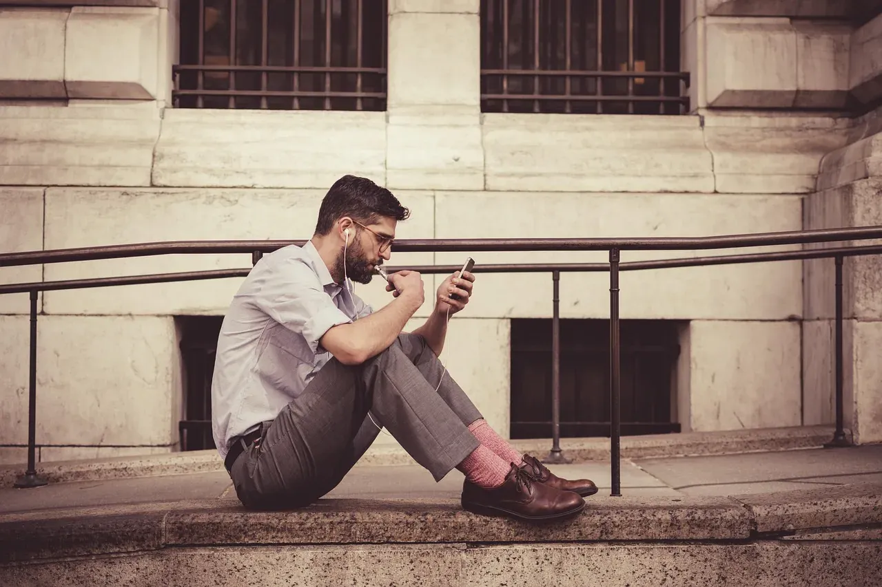 A man sitting against a wall, checking his smartphone