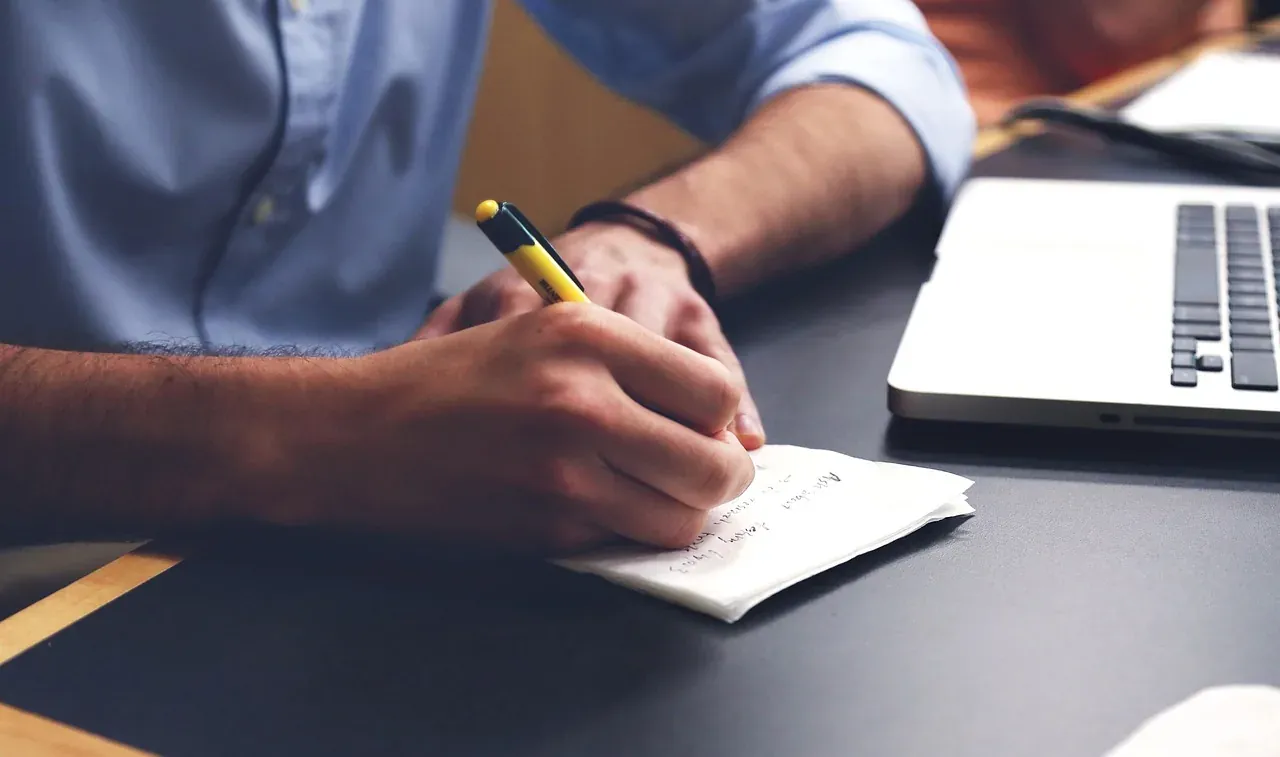 A man writing notes by hand beside a laptop