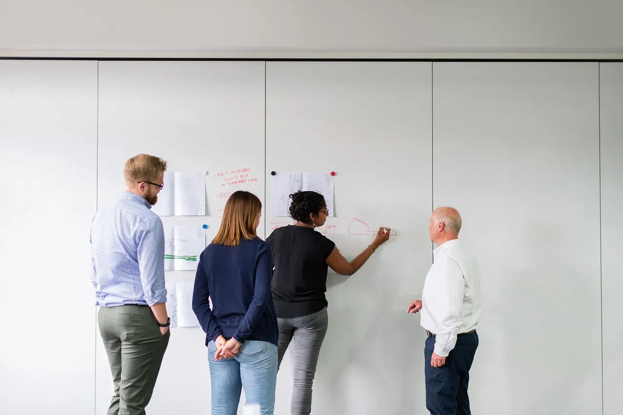 Four colleagues watching as one of them sketches a diagram on an office whiteboard
